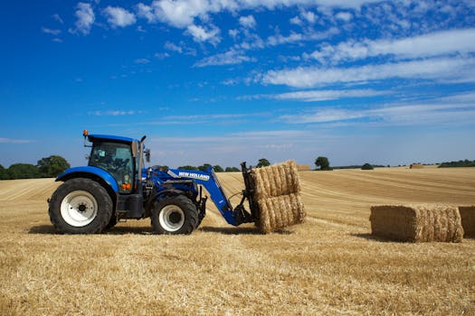 Blue tractor collecting hay bales in a rural English field on a sunny day.